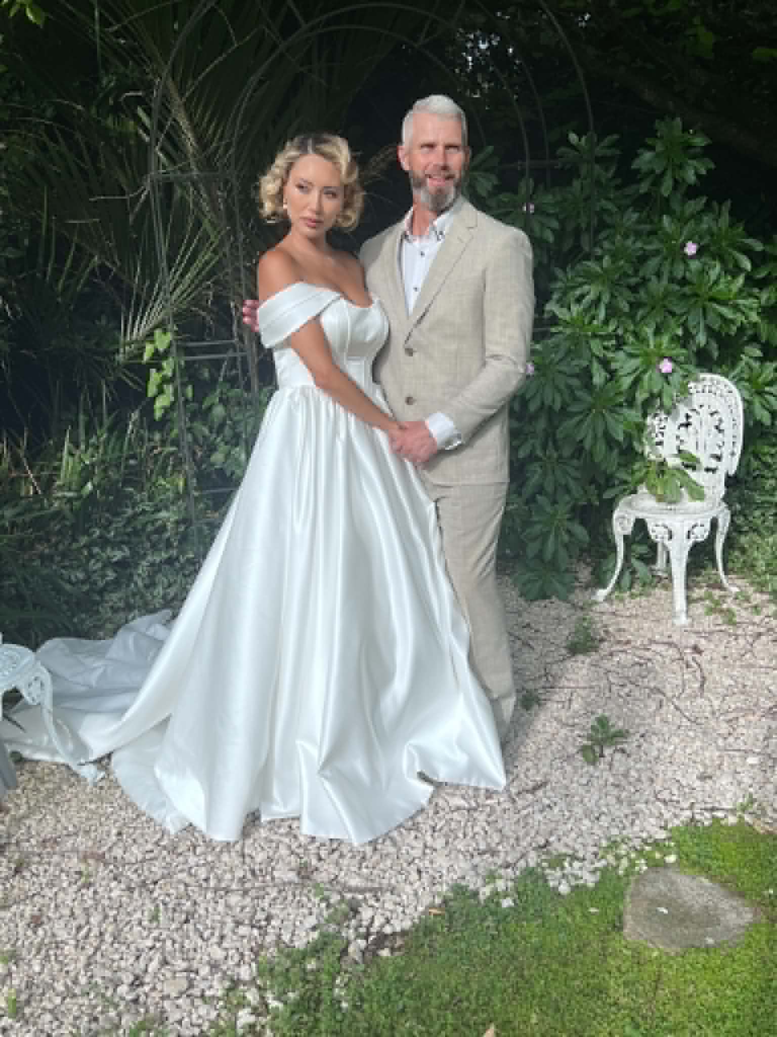 Bride and groom sharing a portrait beneath a garden archway, off-shoulder gown with soft curled updo